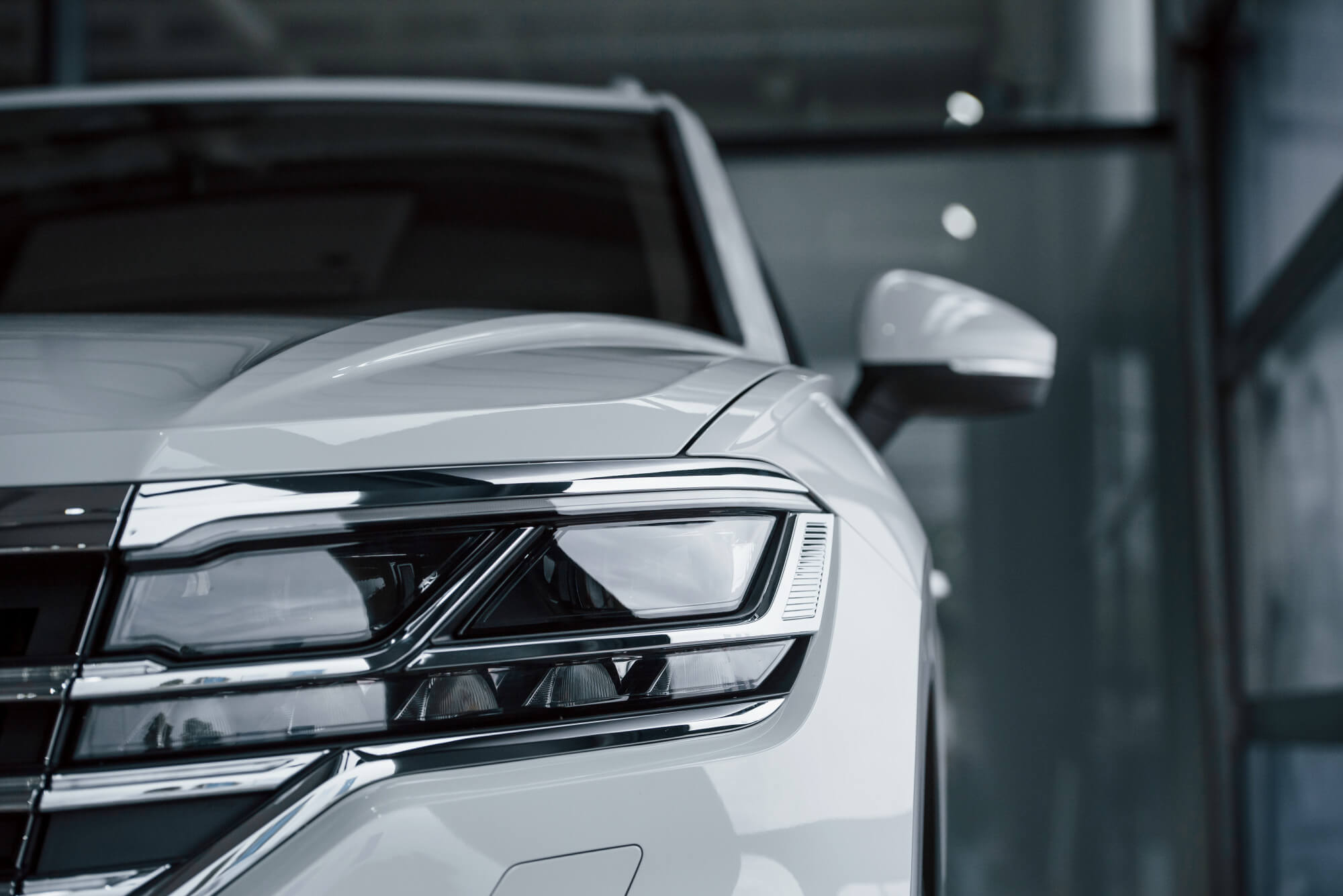 Lähikuva valkoisen auton ajovalosta, jossa on kromattu kehys ja tummat linssit, sekä auton sivupeili ja osa auton korista.